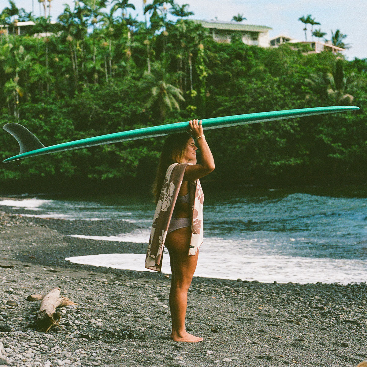 Teal longboard surfboard with quick-dry travel towel on a pebble beach.
