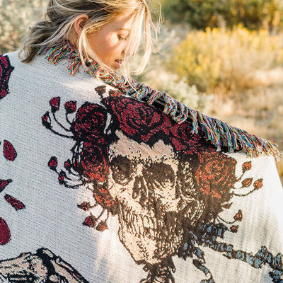 Oversized blanket with skull and red roses pattern, bold fringe, outdoor setting.